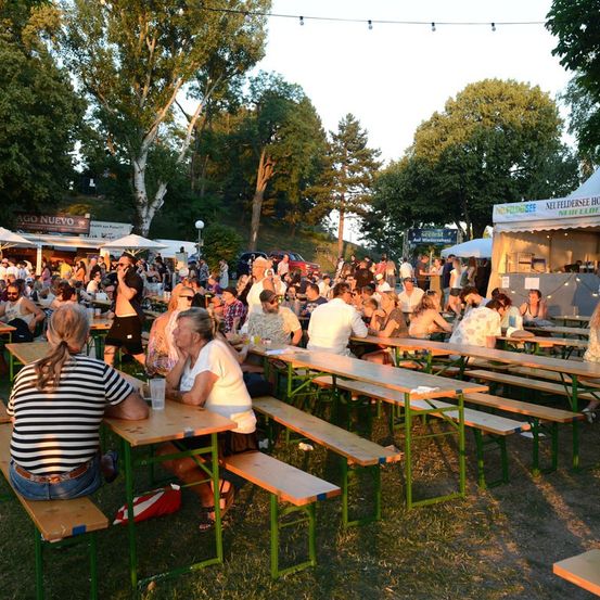 Eine große Gruppe von Menschen versammelt sich bei einer Outdoor-Veranstaltung und sitzt an langen Holztischen mit grünen Metallbeinen. Einige trinken und andere unterhalten sich. Im Hintergrund befinden sich Zelte und Essensstände.