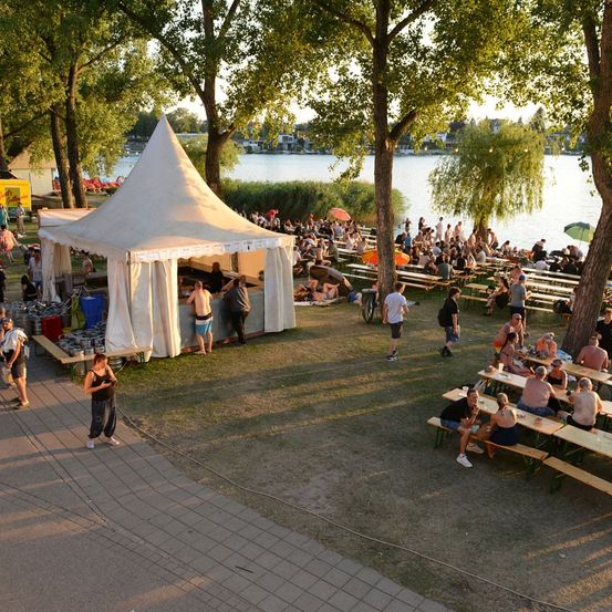 Eine große Outdoor-Versammlung mit vielen Menschen, die an Tischen unter Zelten in der Nähe eines Sees sitzen, einige stehen und gehen herum, und es gibt Bäume und ein gelbes Gebäude im Hintergrund.
