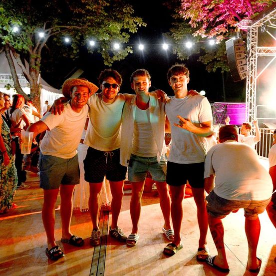 Vier Männer in Freizeitkleidung stehen bei Nacht auf einer Tanzfläche, einer trägt Hut und Sonnenbrille. Hinter ihnen ist eine Menschenmenge zu sehen, und im Hintergrund befinden sich Bäume und Lichter.