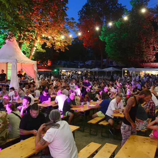 Ein nächtliches Open-Air-Festival mit vielen Menschen, die an Tischen unter bunten Lichtern sitzen, umgeben von Bäumen und Zelten.