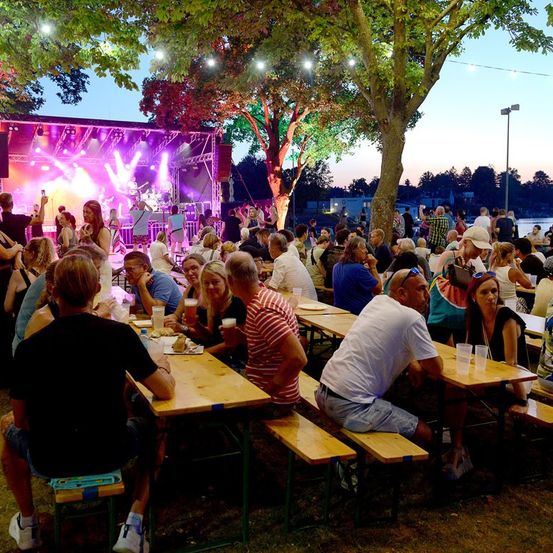 Eine große Menschenmenge versammelt sich um Tische bei einer Outdoor-Veranstaltung, genießt Essen und Getränke und sieht einer Band zu, die auf der Bühne unter bunten Lichtern auftritt.