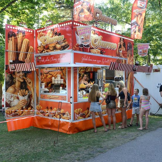 Ein Churro-Stand mit einer Frau im Inneren, die Kunden bedient, eine Gruppe von Menschen, die in der Schlange warten, und ein Banner mit Churros darauf.