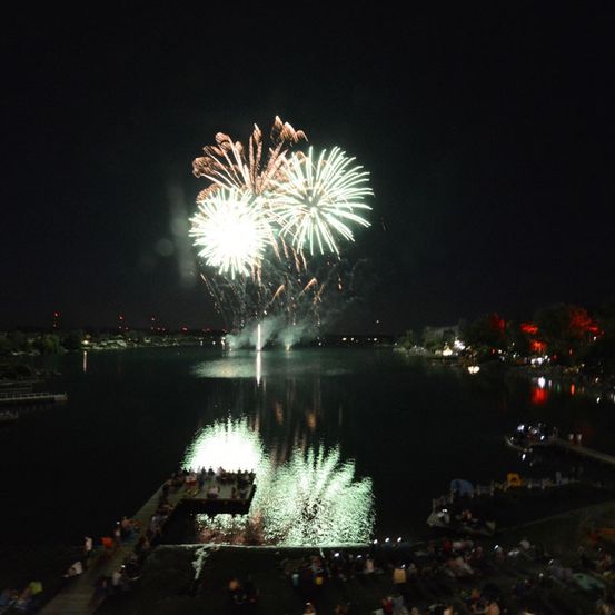 Ein nächtliches Feuerwerk über einem See mit Zuschauern am Ufer und Booten im Wasser.