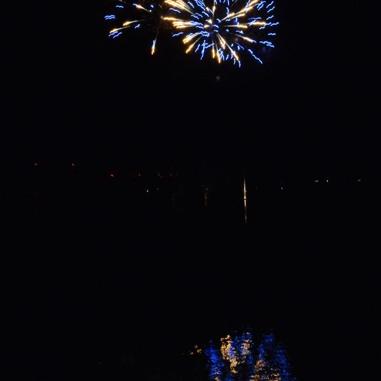 Ein nächtliches Feuerwerk mit blauen und goldenen Explosionen am Himmel, die sich im Wasser darunter spiegeln.