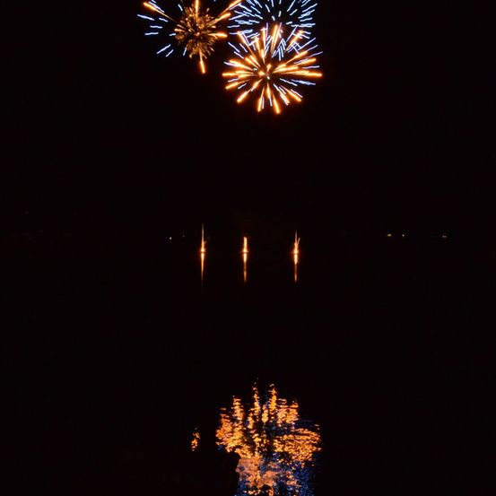 Feuerwerke explodieren in der Nacht über einer ruhigen Wasserfläche, ihre Reflexionen schimmern auf der Oberfläche.