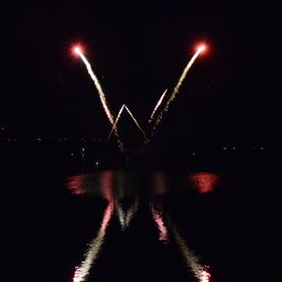 Feuerwerke explodieren in V-Form über einem dunklen Gewässer, wobei die Reflexionen auf der Oberfläche schimmern.