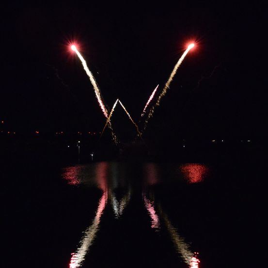Feuerwerke explodieren in V-Form über einem dunklen Gewässer, wobei die Reflexionen auf der Oberfläche schimmern.