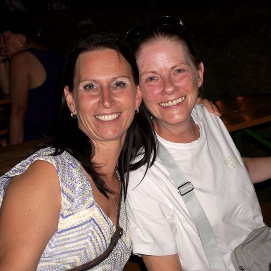 Zwei Frauen lächeln und posieren für ein Foto bei Nacht. Eine hat eine Umhängetasche und die andere hat einen Riemen über der Schulter. Dahinter sitzt eine verschwommene Person auf einer Bank.
