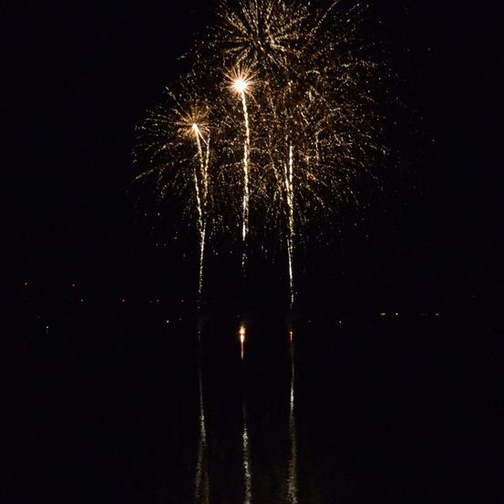 Ein nächtliches Feuerwerk mit drei Lichtblitzen, die den Himmel über einer ruhigen Wasserfläche erhellen.