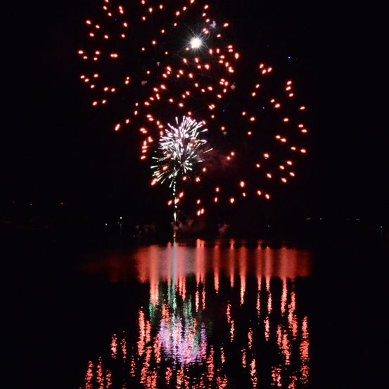 Ein nächtliches Feuerwerk explodiert am Himmel über einer ruhigen Wasserfläche, mit schimmernden Reflexionen auf der Oberfläche.