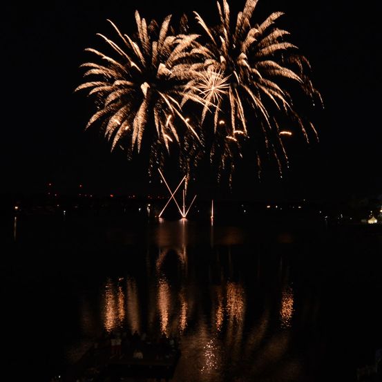 Ein nächtliches Feuerwerk über einem Gewässer, mit Reflexionen auf dem Wasser und entfernten Lichtern im Hintergrund.