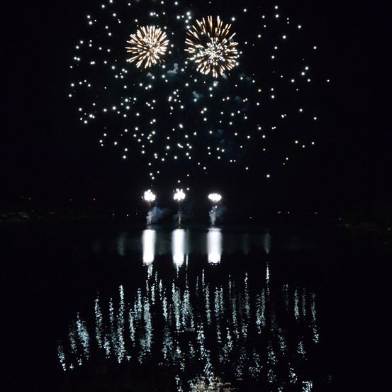 Ein nächtliches Feuerwerk mit zwei großen Explosionen und drei kleineren, die sich im Wasser darunter spiegeln.