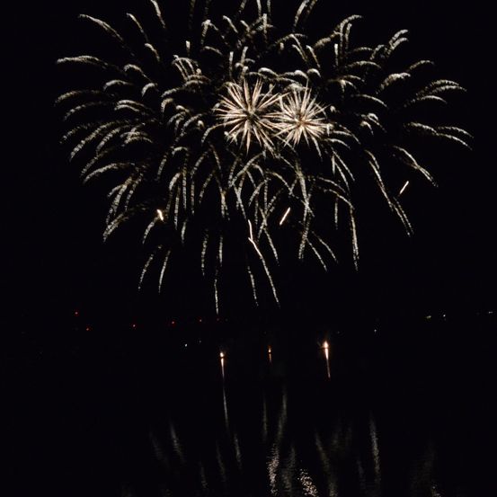 Ein nächtliches Feuerwerk mit mehreren Explosionen, die den Himmel erhellen und sich im Wasser darunter spiegeln.