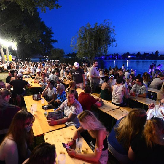Eine große Menschenmenge versammelt sich um Tische bei einer Outdoor-Veranstaltung, wahrscheinlich einem Festival, unter einem Nachthimmel mit Bäumen und einem See im Hintergrund.