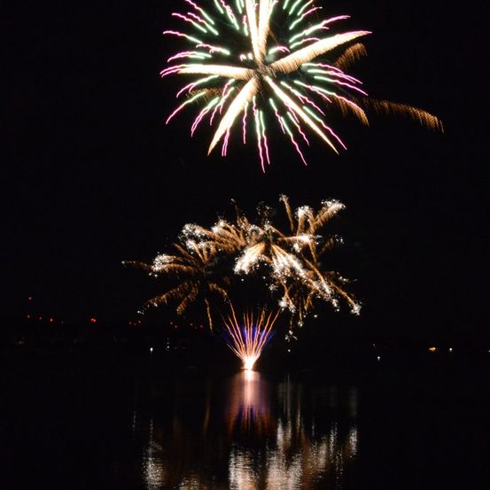 Zwei Feuerwerke explodieren in der Nacht am Himmel, eines in Form einer Blume und eines in Form eines Baumes, die sich im ruhigen Wasser unten spiegeln.