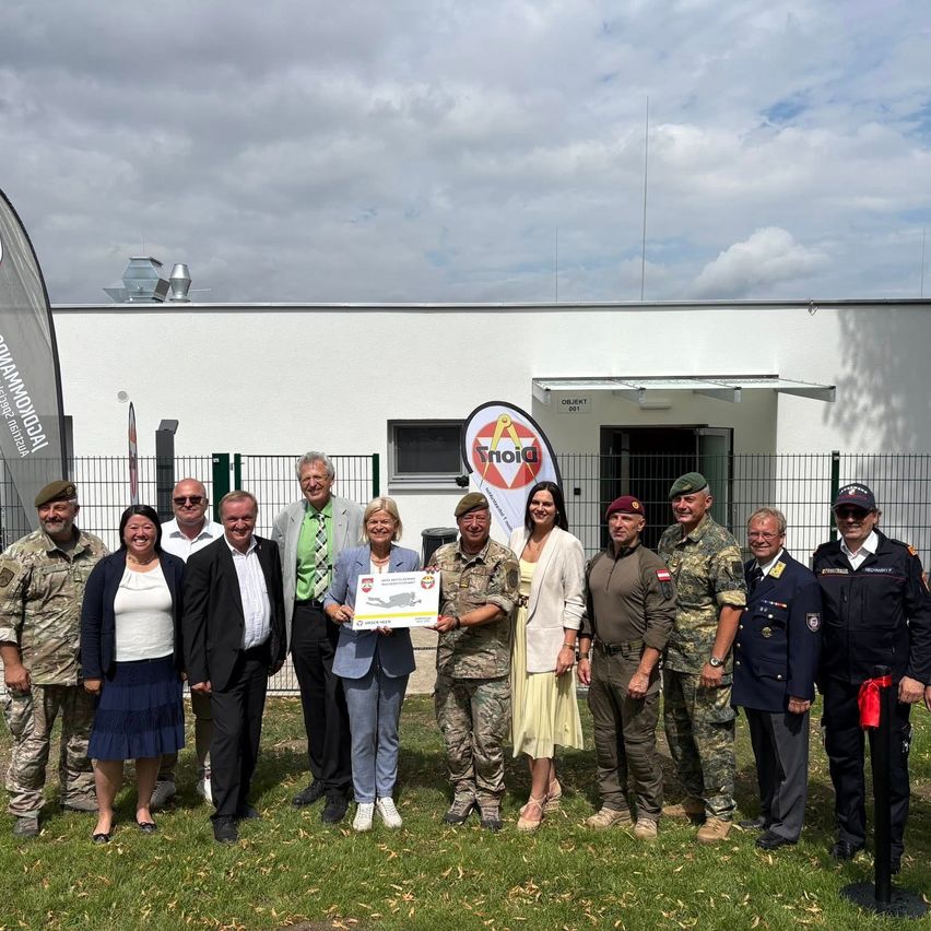 Eine Gruppe von Menschen in Uniformen und Freizeitkleidung steht vor einem Gebäude mit einem Schild, auf dem 'Triola' steht. Sie halten ein Zertifikat und posieren für ein Foto.