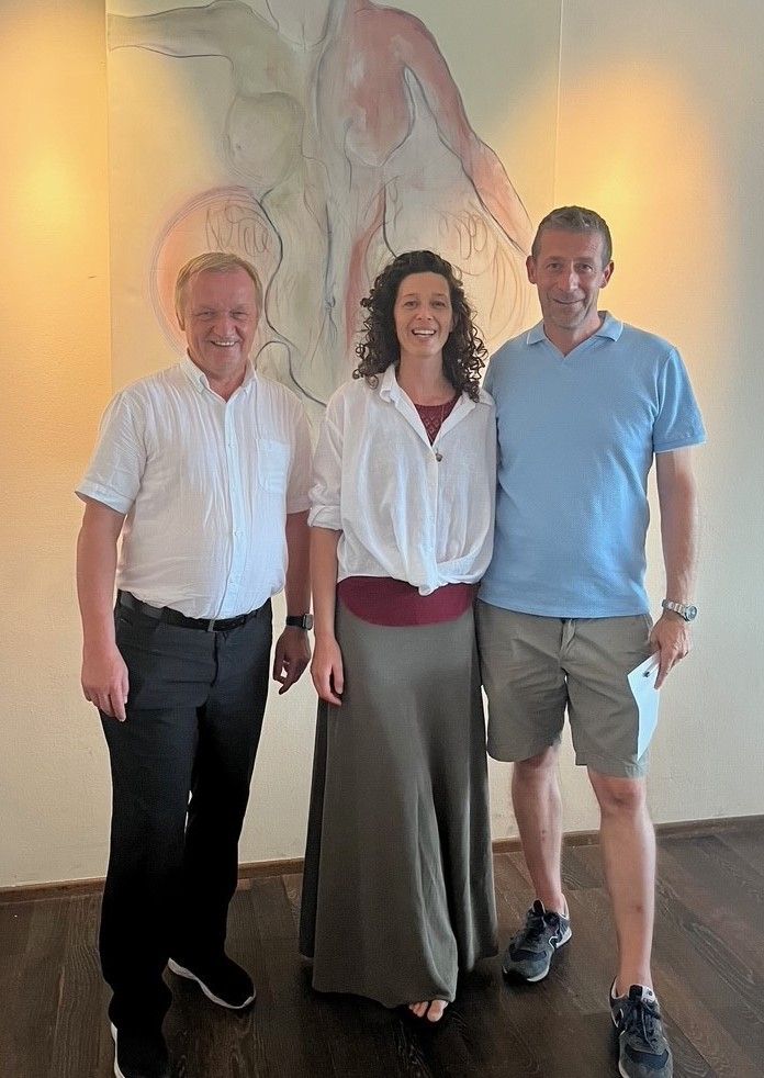 Drei Personen stehen nebeneinander vor einer Wand mit einem Gemälde. Der Mann links trägt ein weißes Hemd und schwarze Hose. Die Frau in der Mitte trägt eine weiße Bluse und einen braunen Rock. Der Mann rechts trägt ein blaues Polohemd und graue Shorts.