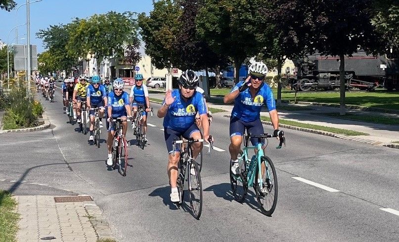 Eine Gruppe von Radfahrern in einheitlichen blauen Trikots fährt eine Straße entlang. Sie tragen Helme und Sonnenbrillen. Bäume und Gebäude säumen die Straße. Ein Zug ist am Straßenrand geparkt.