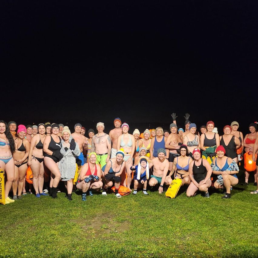 Eine Gruppe von Menschen in Badekleidung und Mützen posiert für ein Foto auf einem Rasenfeld bei Nacht. Viele halten gelbe Taschen.