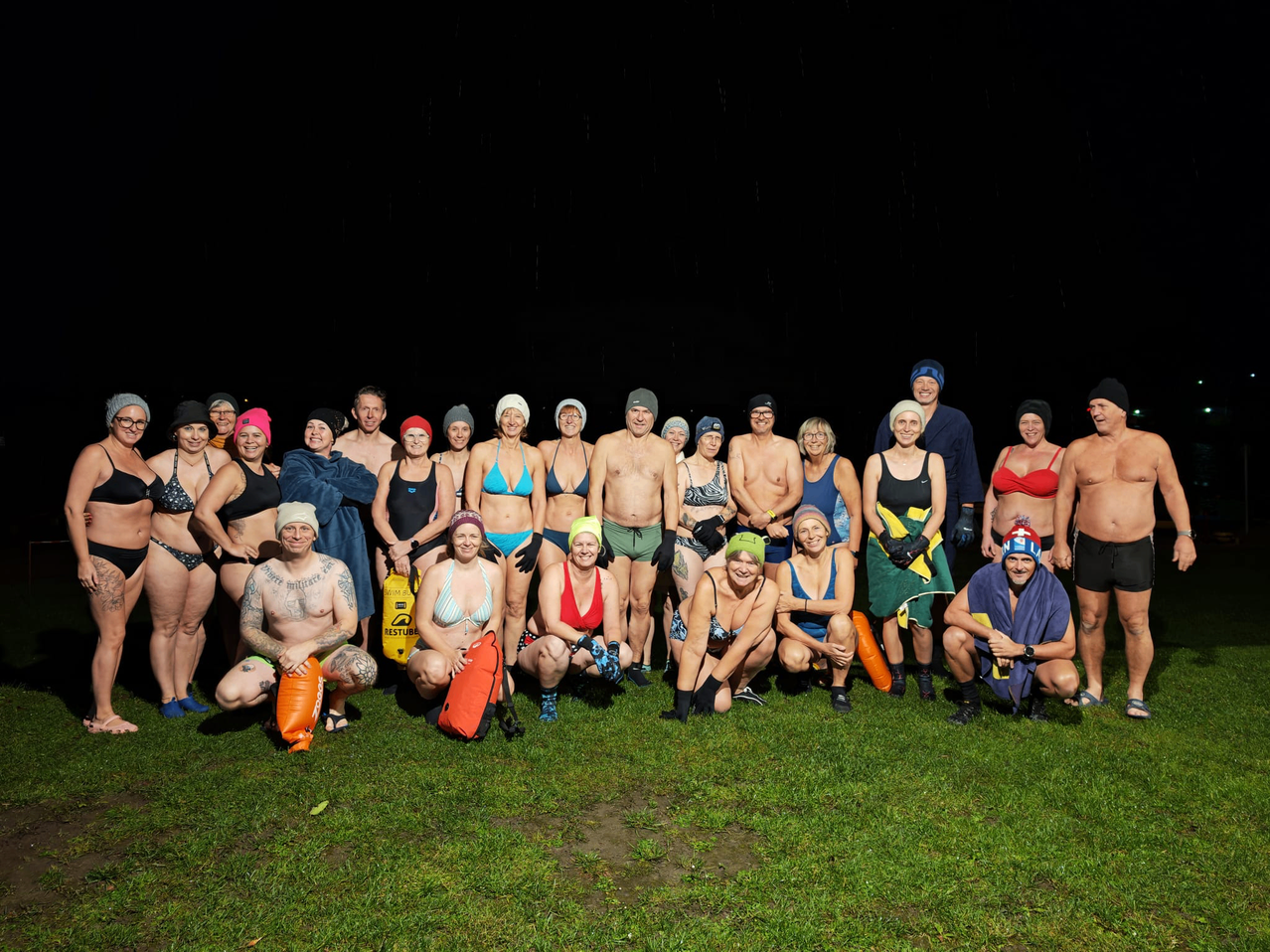 Eine Gruppe von Schwimmern, die meisten in Badekleidung, versammeln sich auf einem Grasfeld bei Nacht, einige halten rote Schwimmhilfen und andere tragen Mützen.