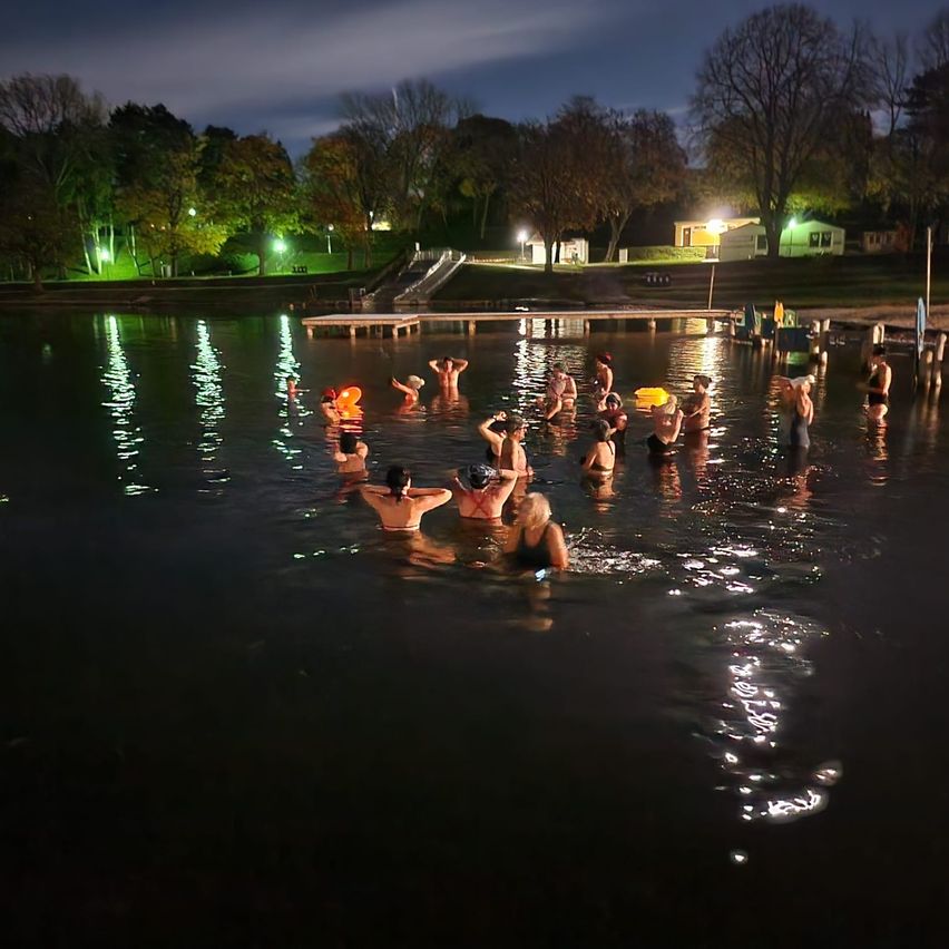 Eine Gruppe von Menschen schwimmt nachts in einem See, der von Lichtern beleuchtet wird. Bäume und Häuser sind im Hintergrund zu sehen.