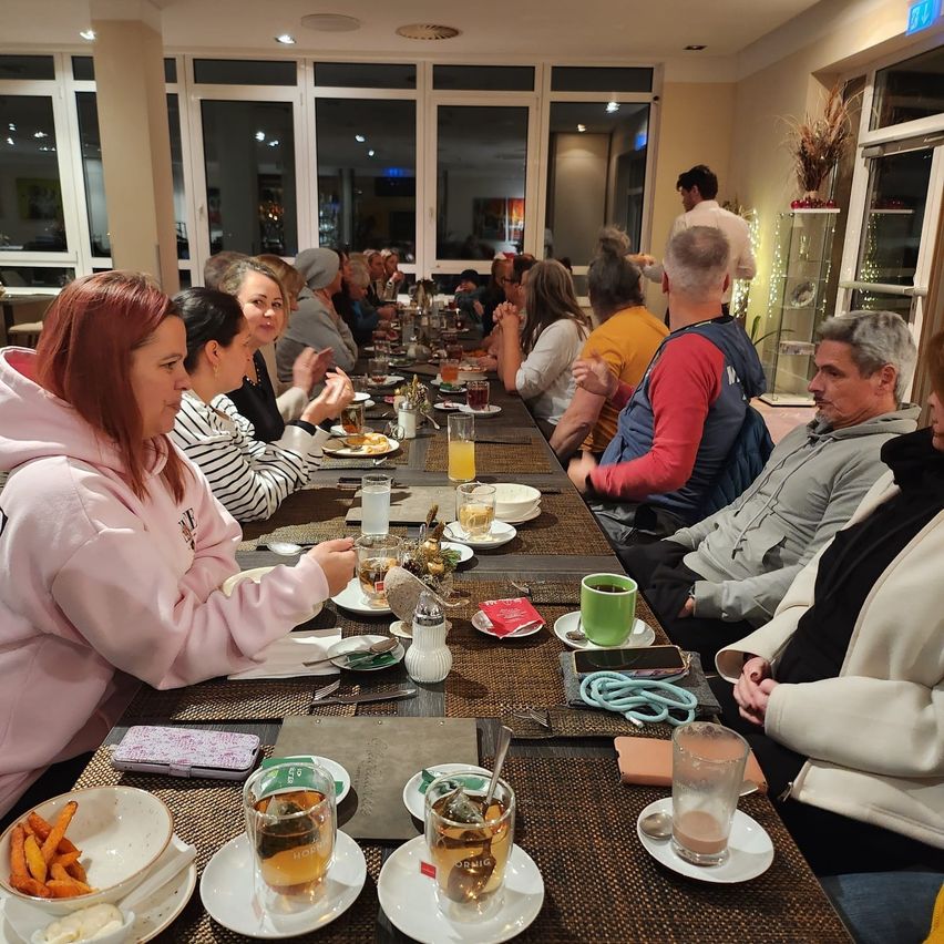 Eine Gruppe von Menschen sitzt an einem langen Tisch, möglicherweise in einem Restaurant. Einige trinken Getränke und essen. Es gibt Teller, Tassen und andere Gegenstände auf dem Tisch. Im Hintergrund befinden sich Glasfenster und eine Tür.