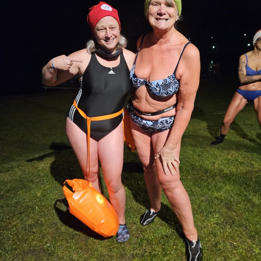 Zwei Frauen stehen auf einem Rasenfeld, eine trägt einen roten Hut und hält eine Tasche, die andere einen blauen Bikini, beide lächeln in die Kamera. Im Hintergrund ist eine weitere Frau teilweise zu sehen.