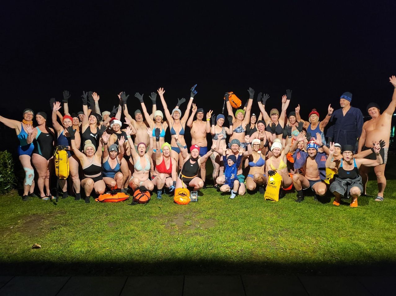 Eine Gruppe von Menschen in Badekleidung, Handschuhen und Mützen posiert nachts auf einem Rasenfeld für ein Foto. Einige heben ihre Hände und tragen Mützen.