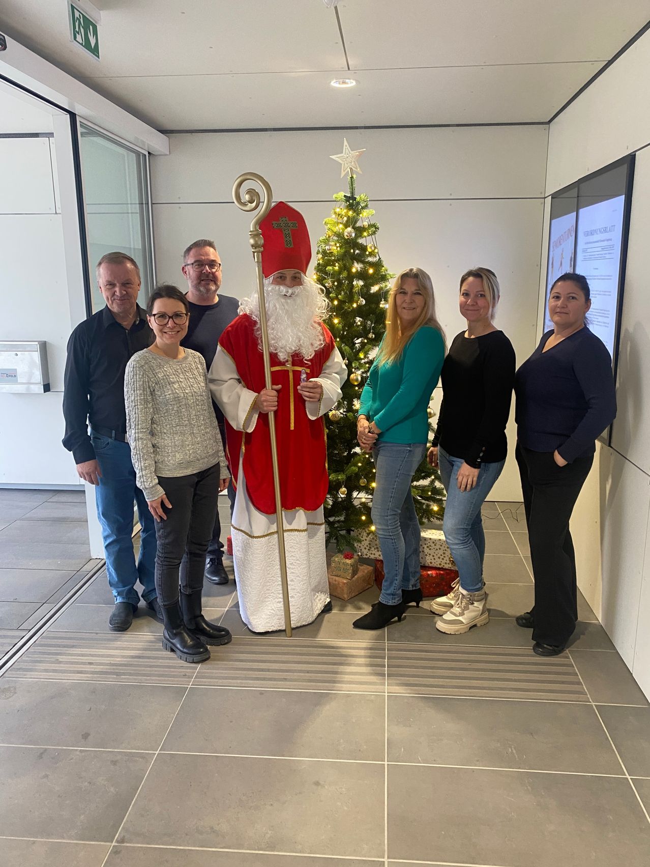 Eine Gruppe von Menschen posiert für ein Foto mit einem Mann, der als Sankt Nikolaus verkleidet ist. Der Mann hält einen Stab und steht vor einem Weihnachtsbaum. Die Leute stehen hinter dem Mann.