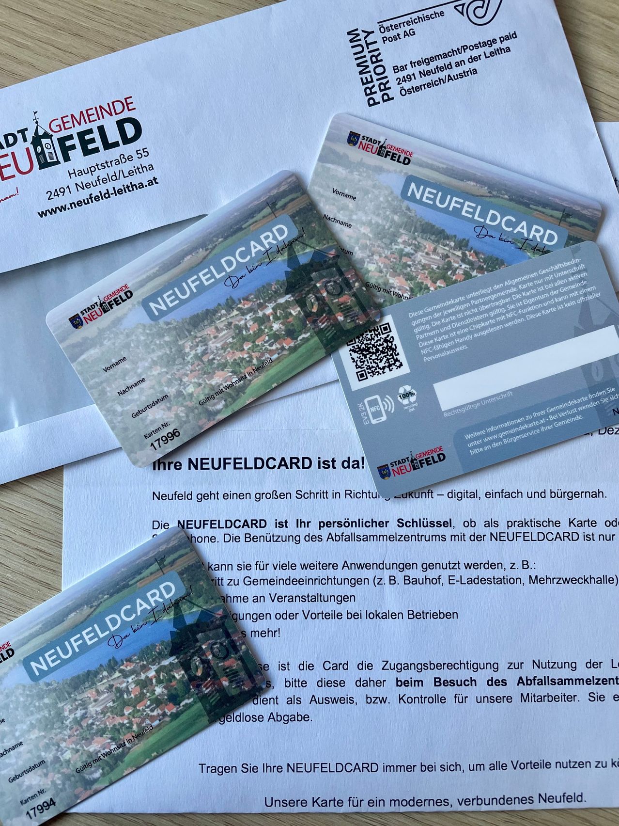 Drei NEUFELDCARD-Karten und ein Brief liegen auf einem Tisch. Die Karten tragen das NEUFELDCARD-Logo und ein landschaftliches Bild. Der Brief informiert über die NEUFELDCARD, einen persönlichen Schlüssel für verschiedene städtische Dienstleistungen.