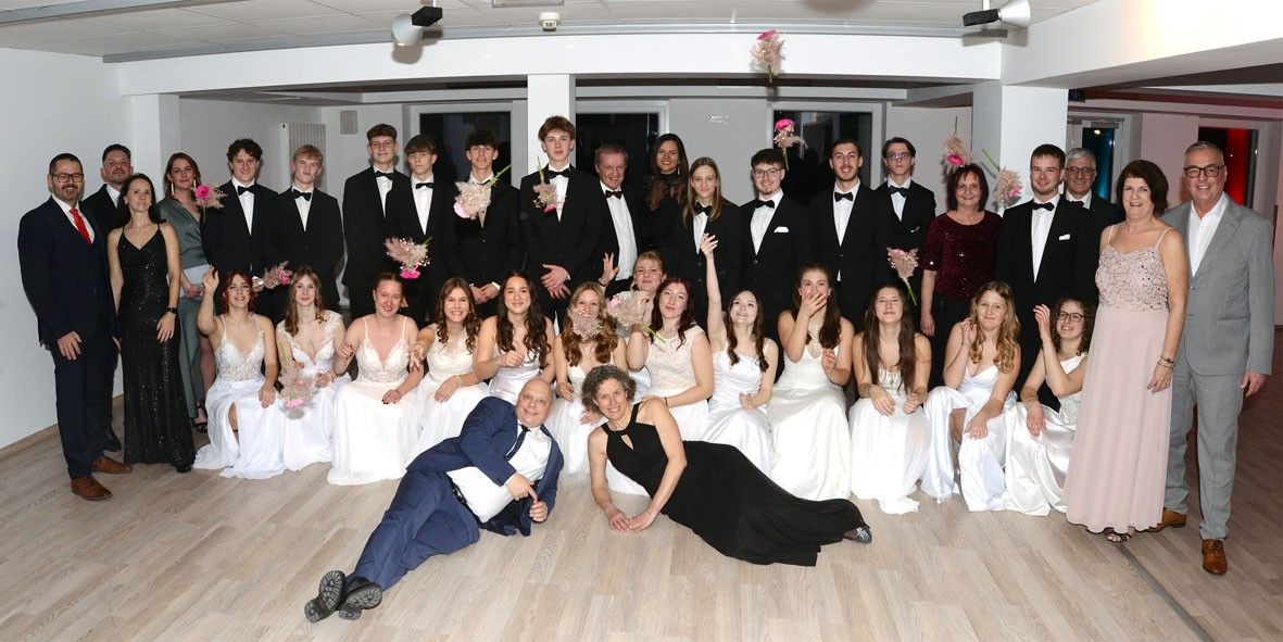 Eine Gruppe von Schülern in formeller Kleidung posiert mit Lehrern für ein Foto in einem Saal.