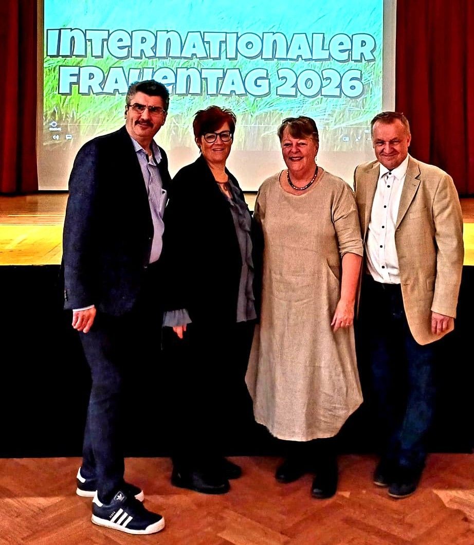 Vier Personen stehen gemeinsam auf einer Bühne und lächeln für ein Foto. Dahinter befindet sich eine Projektionsleinwand mit den Worten 'Internationaler Frauentag 2026'.