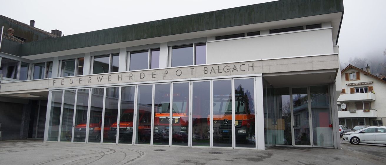 Das Gebäude hat Glaswände und ein großes Schild, das "Uberwehrdepot Balgach" steht. Im Inneren sind mehrere rote Einsatzfahrzeuge geparkt.