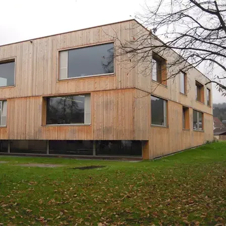 Ein modernes Holzhaus mit vielen Fenstern ist von einem Grasfeld und einem Baum umgeben.