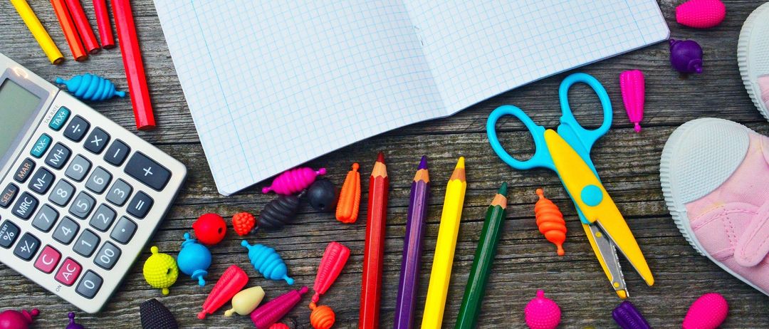 Ein hölzerner Schreibtisch ist mit bunten Schreibwaren bedeckt, darunter ein Taschenrechner, ein offenes Notizbuch, Scheren und verschiedene farbige Stifte und Radiergummis.