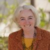 Eine Frau mit weißen Haaren lächelt und posiert für ein Foto. Sie trägt ein gelbes Hemd und eine braune Strickjacke. Sie hat auch eine grüne Halskette. Der Hintergrund ist unscharf.