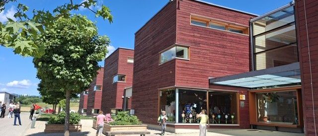 Ein modernes Gebäudekomplex mit roten Fassaden und Glasfenstern. Mehrere Menschen gehen herum und einige stehen in der Nähe der Pflanzen. Es gibt Bäume und Bänke in der Umgebung.