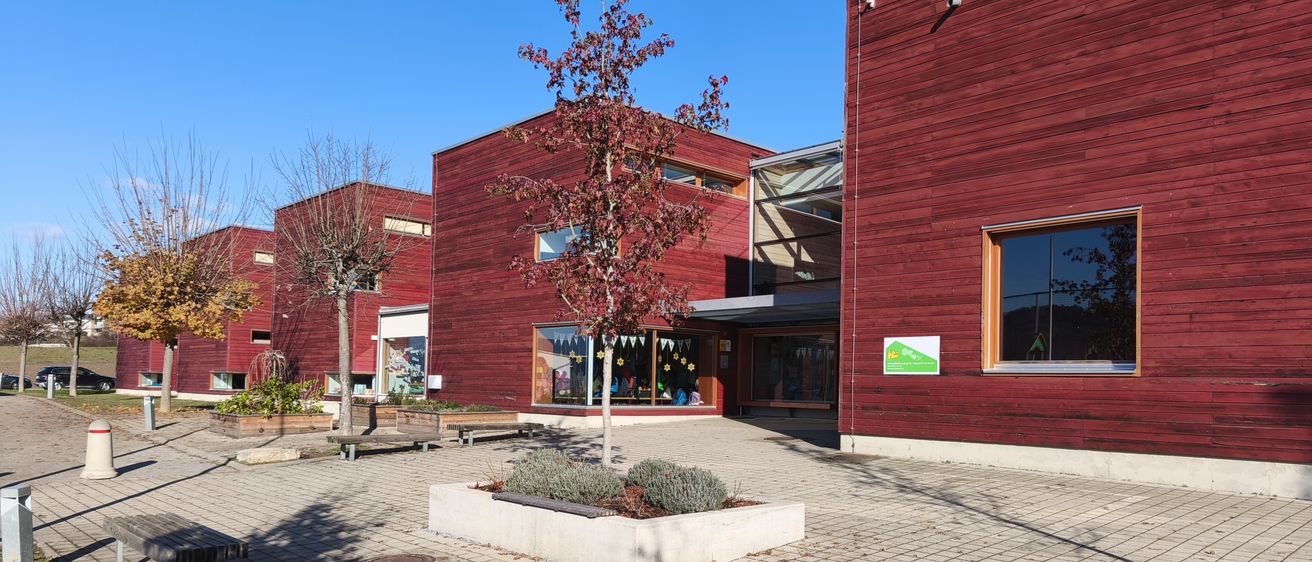 Ein rotes Gebäude mit Holzfassaden und Glasfenstern steht mit einem kleinen Baum in der Mitte eines gepflasterten Bereichs. Bänke sind an den Seiten platziert.