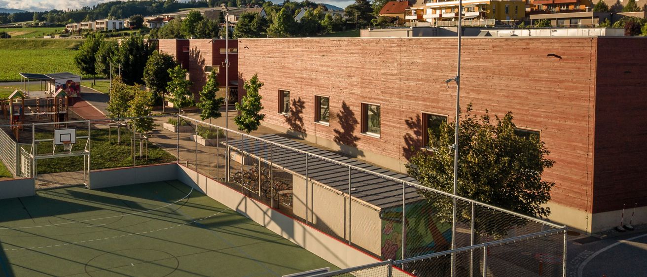 An outdoor basketball court surrounded by a metal fence is shown. Behind it is a building with a brown wall. A playground with trees and grass is also present.