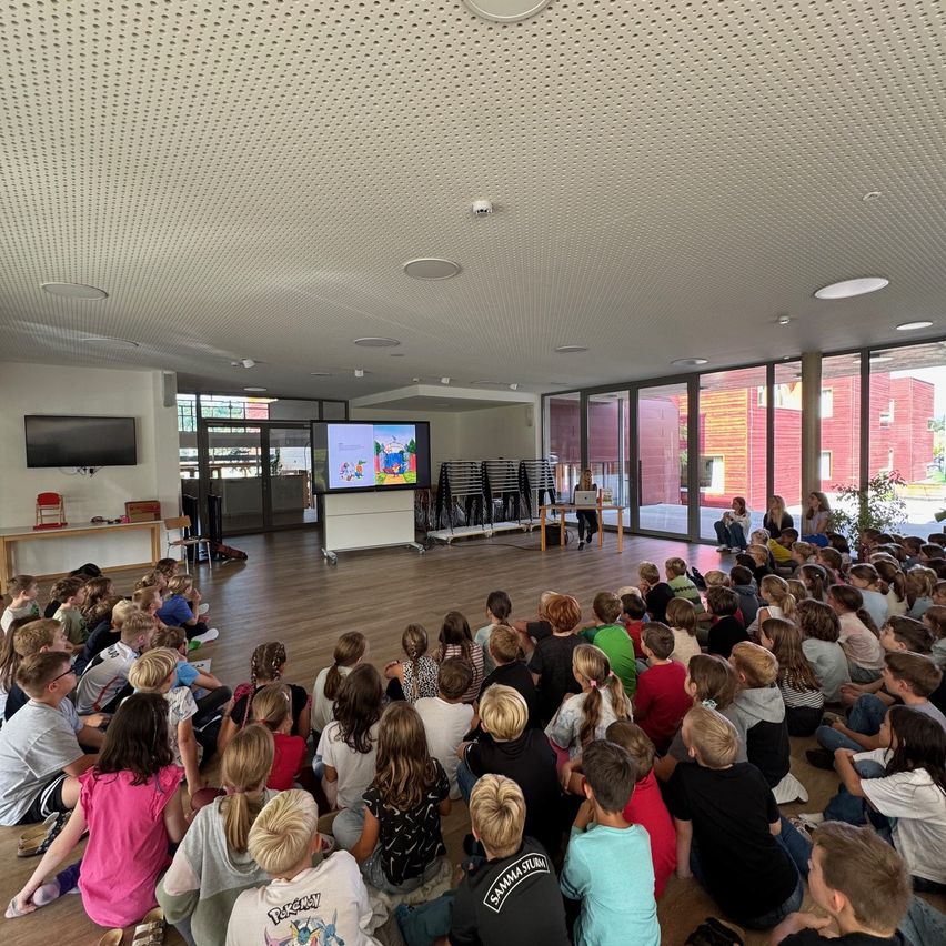 Ein Klassenzimmer mit einem großen Bildschirm, der einen Cartoon zeigt, viele Kinder sitzen auf dem Boden und eine Lehrerin vor einem Schreibtisch.