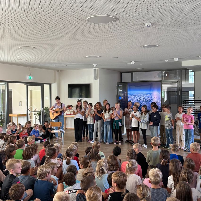 Eine Gruppe von Kindern und einer Frau mit einer Gitarre versammeln sich in einem Raum. Sie führen vor einem großen Publikum auf. Der Raum hat eine Glaswand, einen Fernseher und eine Projektionsleinwand.
