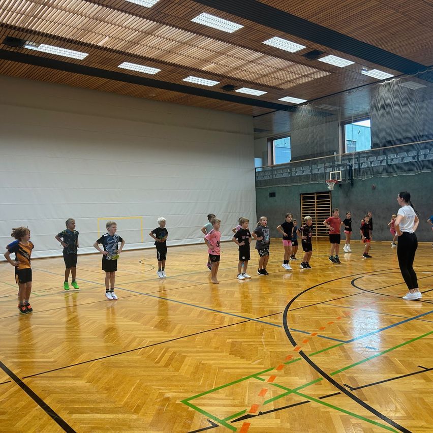 In einer Turnhalle stehen mehrere Kinder in Sportkleidung in einer Reihe, eine Frau steht vor ihnen. Der Boden ist mit Linien markiert und es gibt Fenster im Hintergrund.