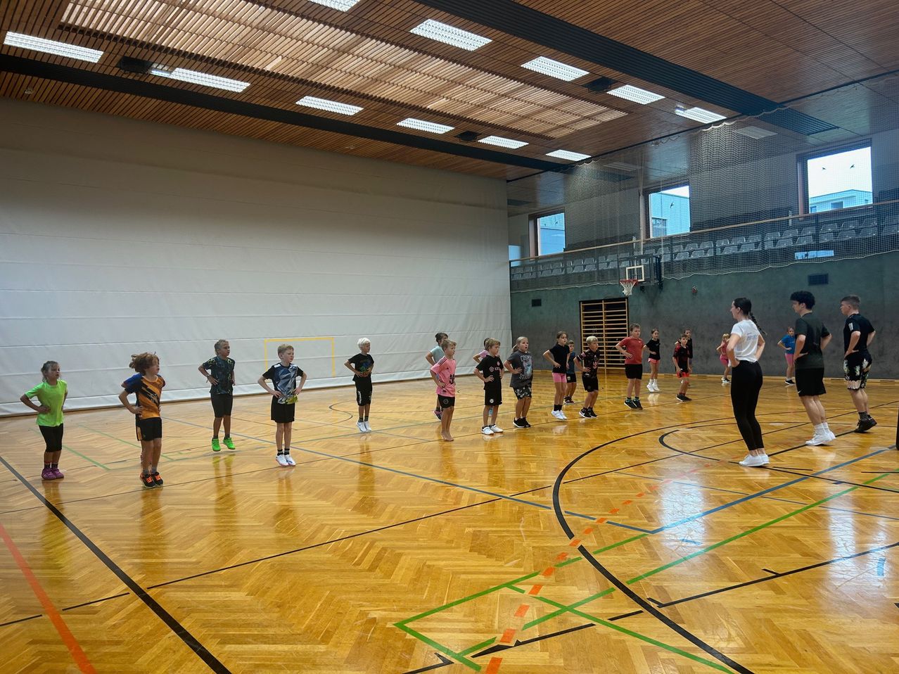 In einer Turnhalle stehen mehrere Kinder in Sportkleidung in einer Reihe, eine Frau steht vor ihnen. Der Boden ist mit Linien markiert und es gibt Fenster im Hintergrund.