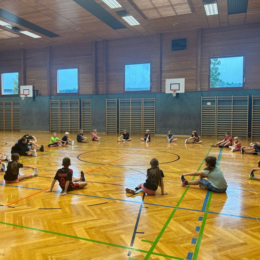 Eine Gruppe von Kindern sitzt auf einem Basketballplatz und ist in ein Spiel verwickelt. Der Platz hat Holzböden und Basketballkörbe.