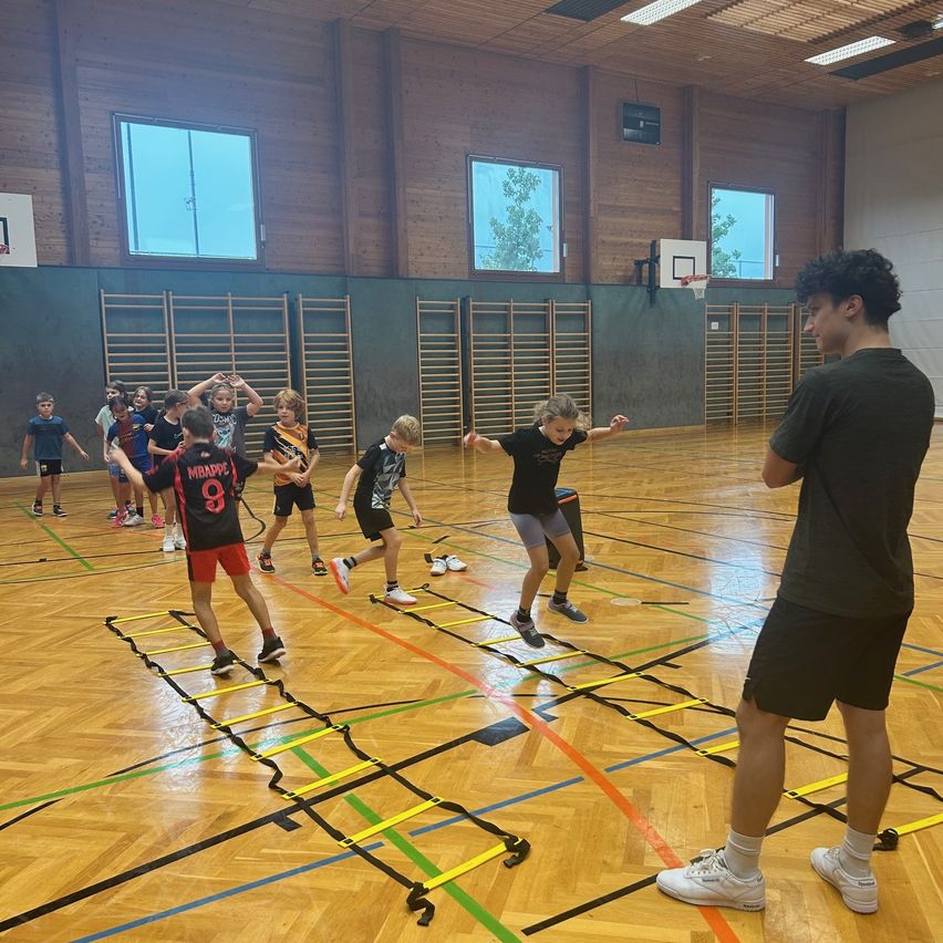 Eine Gruppe von Kindern macht Sprungübungen in einer Turnhalle, wobei ein Trainer beaufsichtigt. Bunte Seile liegen auf dem Boden.