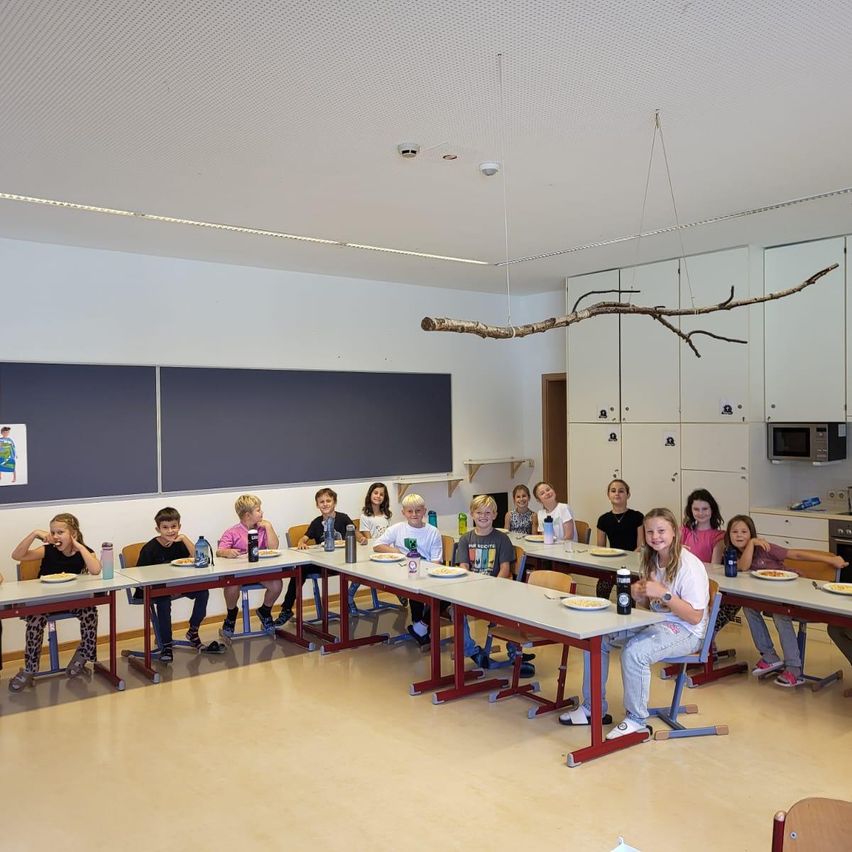 Schüler sitzen an Tischen in einem Klassenzimmer und essen mit Tellern und Flaschen. Eine Tafel hängt an der Wand hinter ihnen, und ein Ast hängt von der Decke.