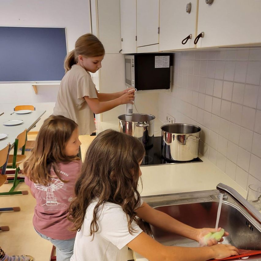 Drei Mädchen sind in einer Küche. Eine wäscht Geschirr, eine kocht und eine beobachtet. Ein Mikrowellenherd, Töpfe und ein Waschbecken sind sichtbar.