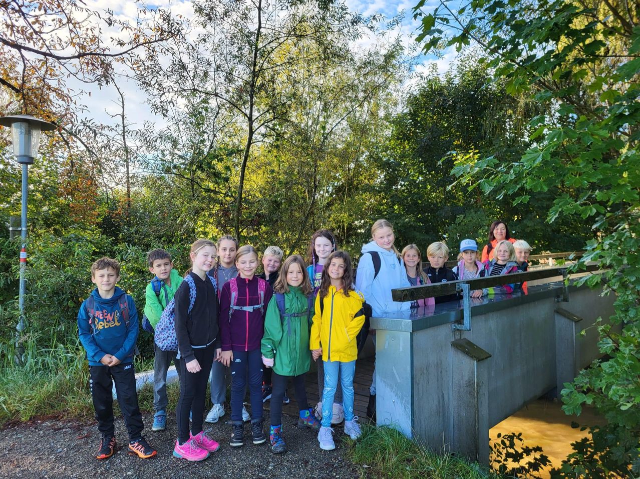 Eine Gruppe von Kindern und einigen Erwachsenen steht auf einer Holzbrücke. Die Kinder tragen Rucksäcke und Turnschuhe. Sie posieren für ein Foto mit Bäumen und einem bewölkten Himmel im Hintergrund.