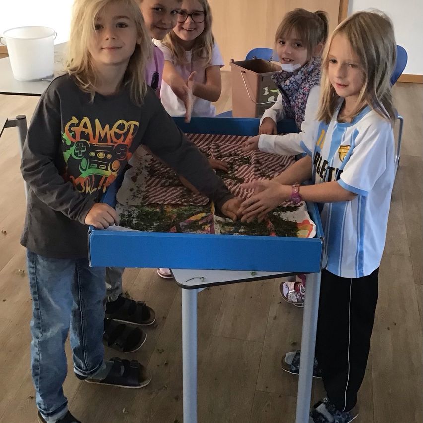 Mehrere junge Kinder stehen um einen blauen Tisch mit grüner Substanz herum. Ein Kind mit einem GAMER-Sweatshirt und eines in einem weißen und blauen Fußballtrikot berühren die grüne Substanz.