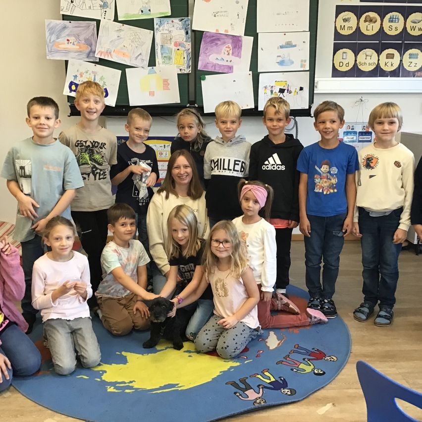 Eine Gruppe von Kindern und eine erwachsene Frau stehen auf einem Weltkartenteppich in einem Klassenzimmer. Einige Kinder knien und halten einen schwarzen Hund.